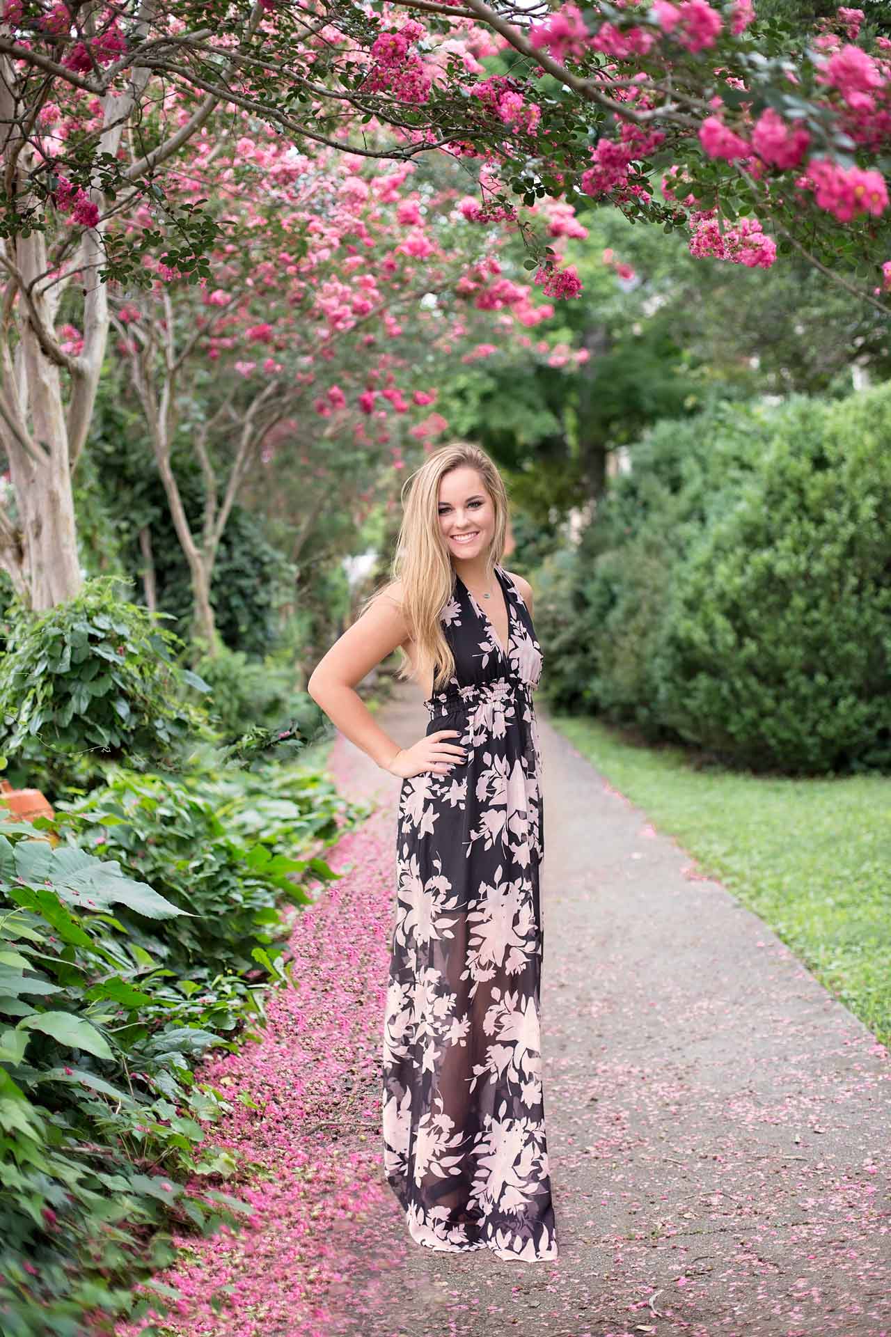 Blonde girl standing on the side walk wearing a black floral dress surrounded by pink crape myrtle trees  during a professional senior photography session at Moments By T, Franklin Tennessee