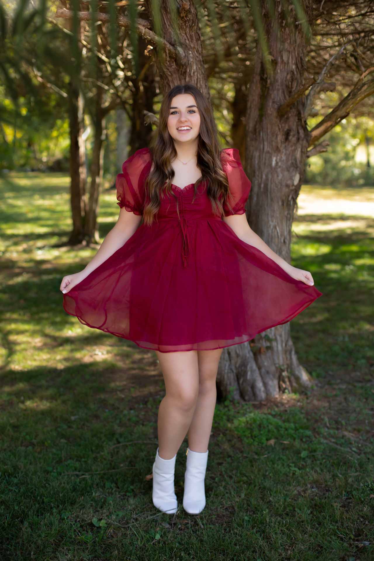 Teen girl standing in a forest wearing a red dress and smiling during a professional senior photography session at Moments By T, Franklin Tennessee