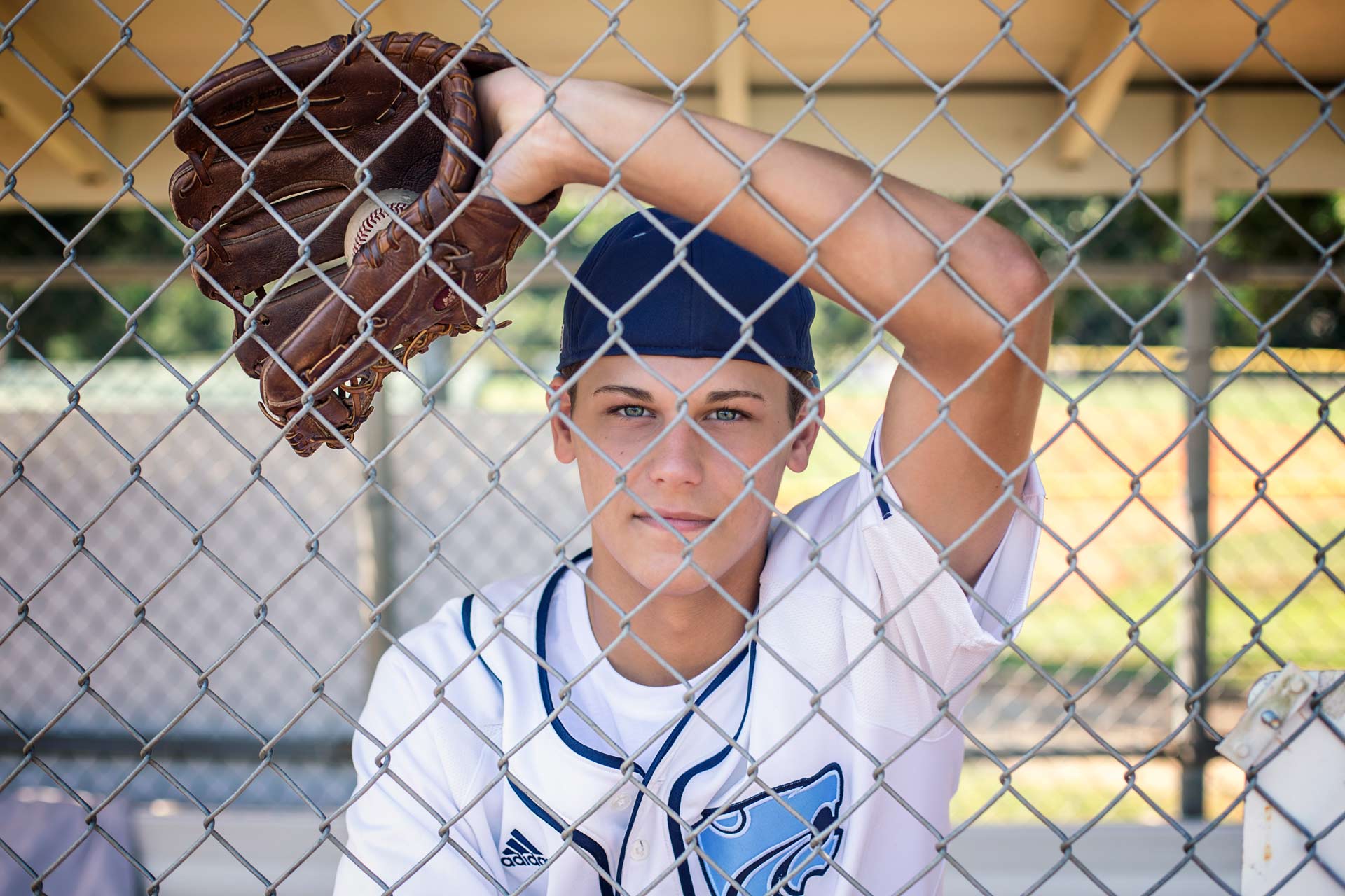 Senior boy standing in the dugout holding a ball in his mitt during a professional senior photography session at Moments By T, Franklin Tennessee