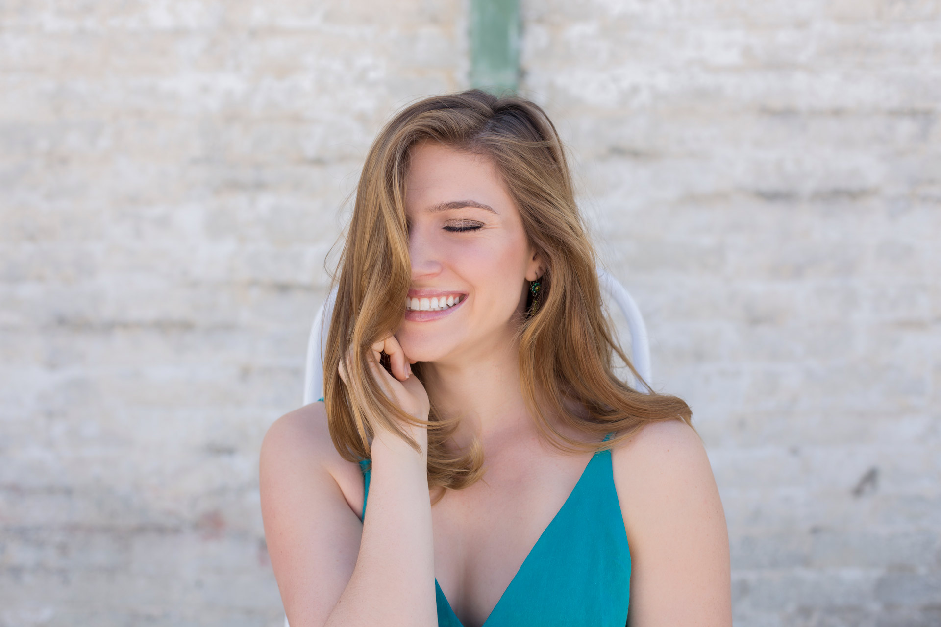 young blonde girl laughing in a green dress in front of a white brick wall. Premier senior portrait photography experience at Harlingdale Farms Franklin TN
